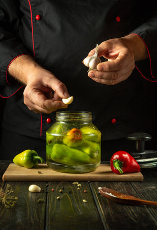 A chef adds garlic to bell pepper before canning them in a jar. Pickling peppers with aromatic spices on the kitchen counter. Advertising space on the black table.の写真素材