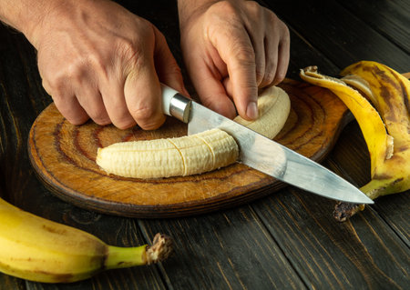 Hands of a cook using a knife on a kitchen board cutting a ripe banana for a diet menu or dish. Bananas diet concept.の写真素材
