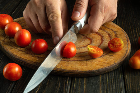 Knife in the cook hand cuts a ripe tomato. Cooking cherry tomatoes on the kitchen table.の写真素材