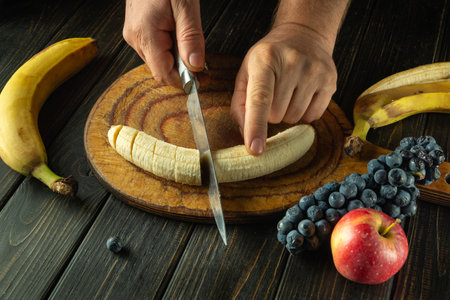 Close-up of chef hands with knife cutting banana before preparing fruit dessert on kitchen table. Concept of preparing healthy dinner with fruits.の写真素材