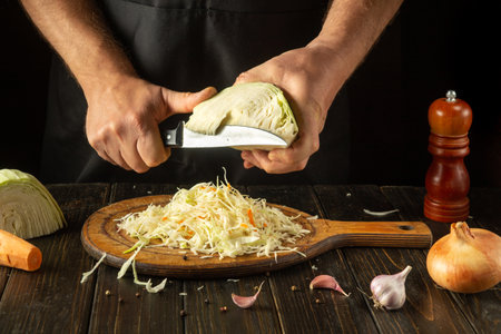 A pair of hands skillfully slices cabbage on a wooden cutting board, surrounded by fresh vegetables like carrots, onions, and garlic, setting the scene for a culinary creation.の写真素材