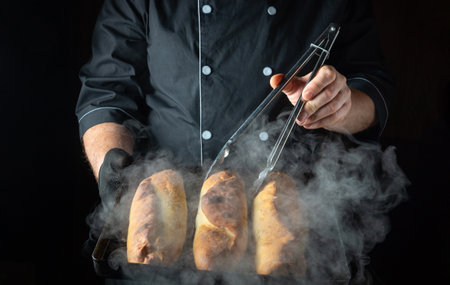 In a warm kitchen filled with steam, a chef uses tongs to lift golden-brown loaves from a baking tray, showing the art of bread-making and culinary expertise in action.の写真素材