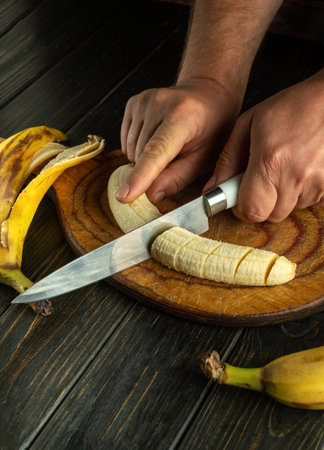 Hands skillfully slice a ripe banana on a rustic wooden board, with fresh bananas nearby. The warm kitchen atmosphere highlights the simple joy of preparing fruit for a tasty snack.の写真素材