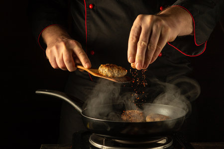A culinary expert skillfully seasons a sizzling dish in a dark kitchen, as aromatic steam fills the air. The careful addition of spices promises a delightful explosion of flavor.の写真素材