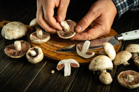 Hands deftly prepare fresh mushrooms on a wooden cutting board, showcasing the artistry of cooking in a warm kitchen environment. A knife glints beside them, ready for action.の写真素材