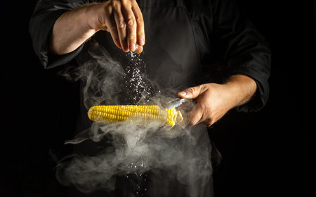 A skilled chef adds seasoning to freshly grilled corn on the cob, creating a captivating display of spices. Wisps of smoke swirl around as flavors blend in a dimly lit kitchen.の写真素材