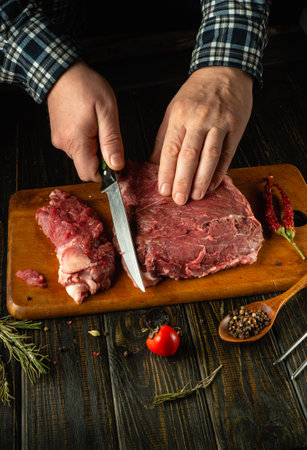 A pair of skilled hands slice a large piece of raw meat on a wooden cutting board, surrounded by fresh herbs and spices.の写真素材