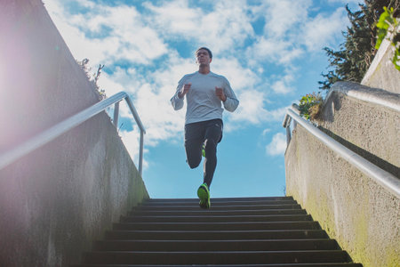 A fit man jogs up a set of outdoor stairs, surrounded by lush greenery and a blue sky. The sun shines brightly, casting playful shadows as he embraces his fitness journey.の素材
