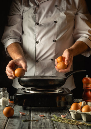 Hands expertly hold fresh eggs above a hot skillet, ready for cooking. The ambiance is warm and inviting, with kitchen tools and ingredients arranged nearby, enhancing the culinary atmosphere.の写真素材