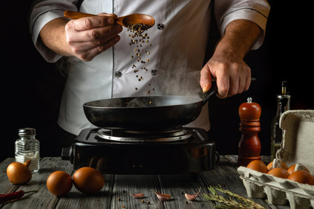 A skilled chef adds fresh peppercorns to a hot pan, releasing aromatic steam. The kitchen is lively with ingredients like eggs, garlic, and herbs, creating a delicious culinary atmosphere.の写真素材
