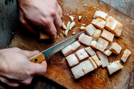 Hands engage in a culinary activity, chopping fresh pork fat and garlic on a rustic wooden board. The kitchen exudes an inviting atmosphere, perfect for cooking enthusiasts.の素材