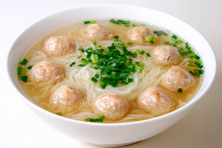 A steaming bowl of noodle soup fills the table, showing delicate strands of noodles paired with savory meatballs. Fresh herbs add a vibrant touch, inviting a warm dining experience.の素材