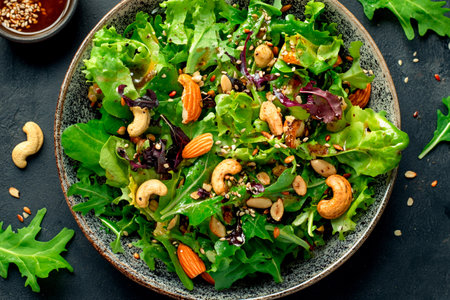 A colorful salad made with fresh mixed greens, cashews, almonds, and a sprinkle of seeds sits elegantly in a bowl beside a small dish of dressing.の素材