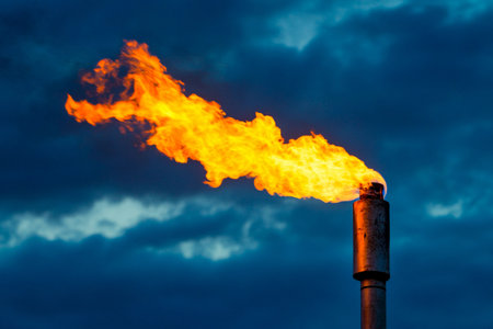 Bright orange flames erupt from a metal flare stack, illuminated against a backdrop of dark, swirling clouds at dusk. The contrasting colors create a striking visual impact.の素材