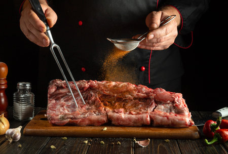 A chef meticulously seasons a large cut of meat using a strainer and a fork. Fresh herbs and spices are prominently present on the wooden board.の写真素材