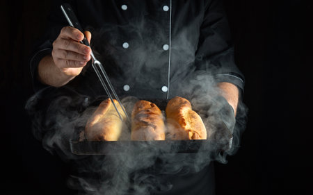 Fork and baking tray with hot buns in the hands of an experienced baker. Low-key concept of making fresh and delicious buns.の写真素材