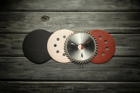 A collection of sanding discs in various colors and a circular saw blade are neatly arranged on a rustic dark wood surface. The tools highlight woodworking equipment used for shaping and finishing.の写真素材