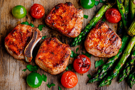 Succulent grilled pork chops are arranged on a rustic wooden table alongside bright green tomatoes, cherry tomatoes, and fresh asparagus, showing a vibrant meal ready to enjoy.の素材