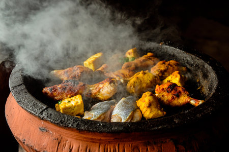 Grilling takes place on a clay grill with various meats, including chicken and fish, surrounded by billowing smoke. This outdoor preparation showcases vibrant colors and hearty flavors.の素材