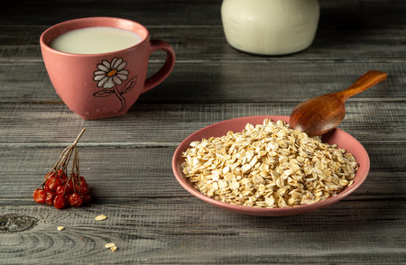 A bowl of rolled oats sits beside a pink mug filled with milk. A wooden spoon is placed in the bowl, while red berries add a pop of color to the rustic setting.の写真素材