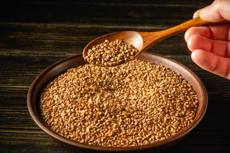 A hand holds a wooden spoon over a bowl filled with buckwheat grains on a dark wooden surface. The scene conveys the texture and color of the grains, highlighting their natural appearance.の写真素材