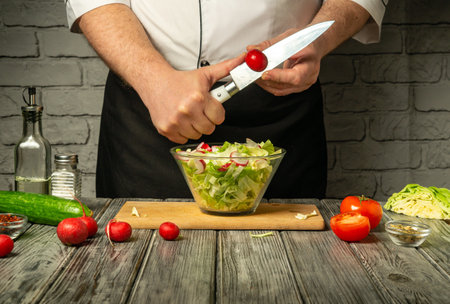 A chef skillfully chops vegetables while preparing a fresh salad. Ingredients on the wooden table include lettuce, cucumbers, tomatoes, and radishes, showcasing a vibrant kitchen atmosphere.の写真素材