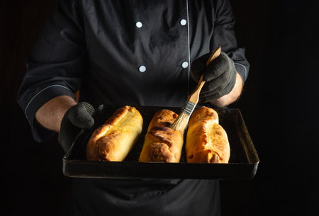 A skilled chef holds a tray of freshly baked goods, brushing them with glaze. The kitchen setting is dimly lit, emphasizing the warm, inviting colors of the pastries.の写真素材