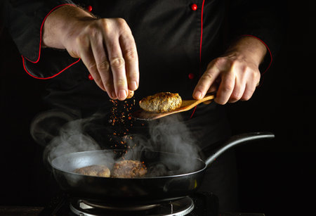 A chef skillfully adds spices to sizzling meat patties in a frying pan, producing flavorful aromas in a kitchen filled with steam during a nighttime cooking session.の写真素材