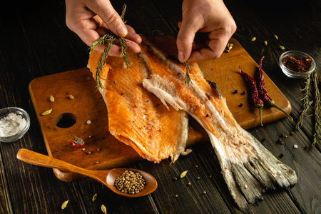 Hands are carefully seasoning a fresh salmon fillet with herbs and spices. The wooden cutting board, salt, and dried peppers add a rustic charm to the cozy kitchen atmosphere.の写真素材
