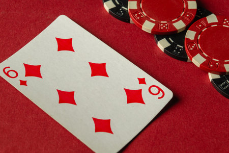 A close-up view of a playing card showing the six of diamonds besides black and red poker chips. The table is covered in vibrant red felt as players engage in a lively game.の写真素材