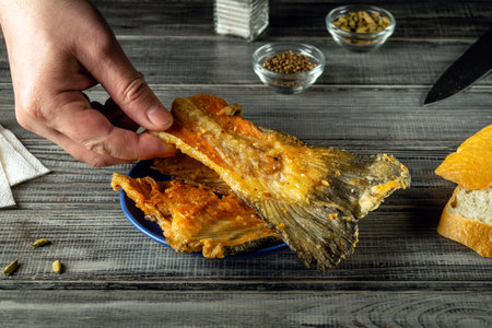 A hand lifts a piece of crispy fish from a blue plate filled with more fish beside slices of bread...