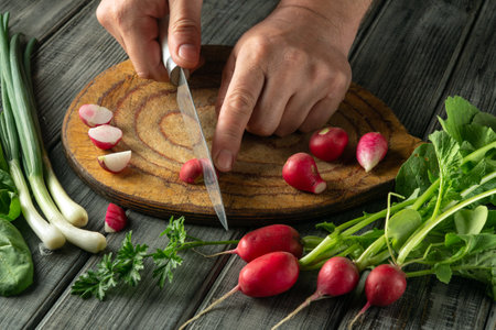 A person is chopping fresh radishes on a wooden cutting board in a kitchen. Surrounding vegetables include green onions and leafy greens, suggesting meal preparation.の写真素材