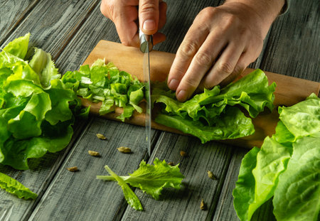 Fresh lettuce is being chopped into smaller pieces on a wooden cutting board. The vibrant green leaves are arranged neatly. Natural light brightens the kitchen atmosphere.の写真素材