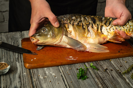 A person's hands hold a freshwater fish on a wooden cutting board while preparing to fillet it. Nearby, various herbs and spices are visible, creating a culinary atmosphere.の写真素材