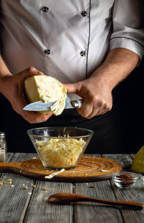 A chef skillfully shreds a head of cabbage with a knife, placing the freshly cut pieces into a transparent bowl. The kitchen setup features rustic wooden elements and spices.の写真素材