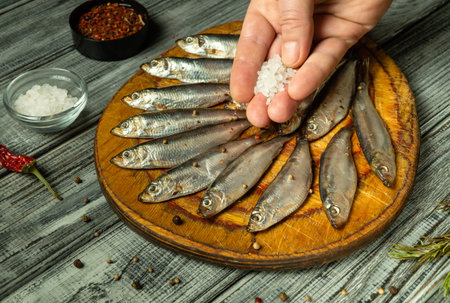 A hand sprinkles coarse salt over fresh small fish arranged neatly on a wooden cutting board. Various spices are scattered around, enhancing the cooking atmosphere.の写真素材