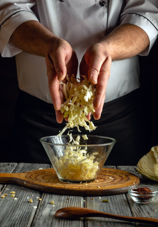 A skilled cook is grating fresh cabbage into a glass bowl on a wooden cutting board. The workspace features rustic elements and a warm, inviting atmosphere.の写真素材