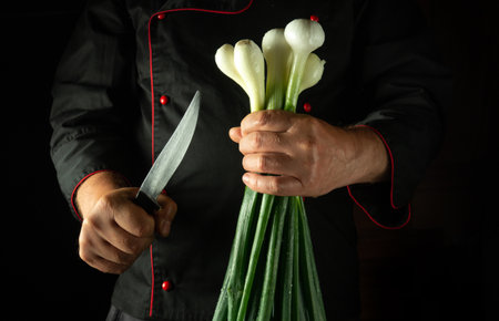 A chef stands in a dimly lit kitchen, gripping a bunch of fresh green onions in one hand while...