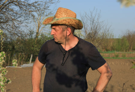 A man wearing a straw hat stands in a garden, looking thoughtfully at the plants. The sun is shining and the atmosphere is calm in this rural landscape.の写真素材