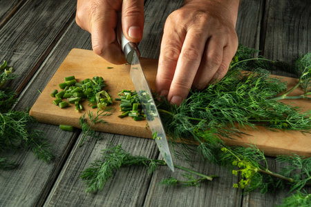 Fresh green herbs are being chopped with a sharp knife on a wooden cutting board. The hands show a precise technique as sunlight filters into the kitchen, enhancing the vibrant colors.の写真素材