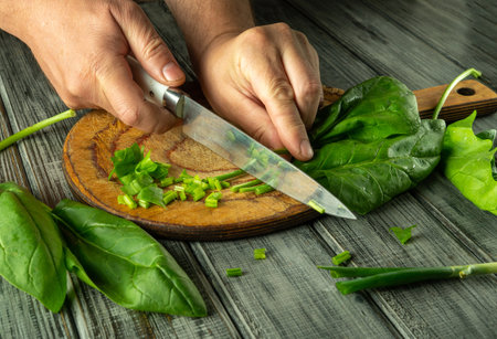 Two hands are skillfully chopping green onions and leafy greens on a wooden cutting board in a bright, rustic kitchen. The vibrant colors of the ingredients are highlighted.の写真素材