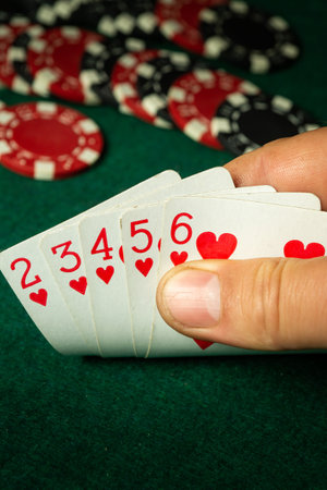 A hand holds five playing cards displaying hearts. Red and black poker chips are arranged on a green...