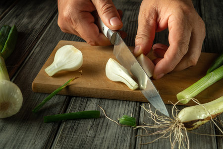 Hands skillfully chop green onions on a wooden cutting board, with vegetables scattered nearby, showcasing a culinary preparation moment in a warm kitchen.の写真素材