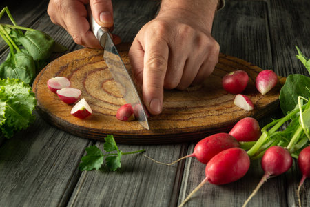 A person skillfully slices fresh radishes on a wooden board. Surrounding the board are vibrant greens, indicating a focus on healthy cooking.の写真素材