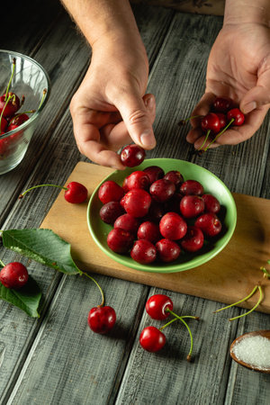 Hands delicately select ripe cherries from a green bowl filled with fruit in a cozy kitchen. The setting features wooden surfaces and scattered fruit, evoking a homemade feel.の写真素材