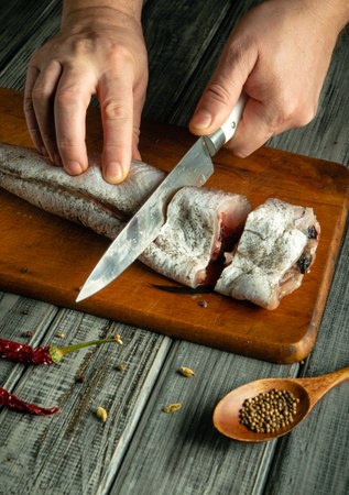 Hands are slicing a fresh fish on a wooden surface. Nearby, dried chilies and a spoon of coriander seeds create a rustic cooking atmosphere in a warm kitchen.の写真素材