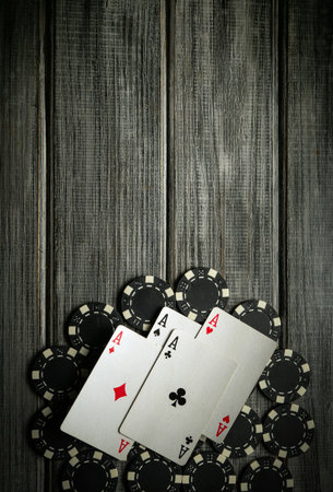 Four Aces playing cards featuring aces are centered among several black poker chips on a weathered wooden table. The setting suggests a relaxed game night atmosphere.の写真素材