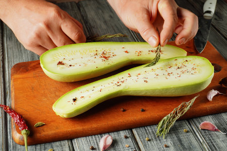 Hands delicately slice open fresh squash, revealing the tender inside, while sprinkling spices and herbs on the wooden cutting board. A knife and garlic cloves accompany the setup.の写真素材