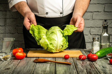 In a rustic kitchen, a chef holds a large, fresh cabbage above a wooden cutting board surrounded by colorful vegetables. Natural light illuminates the workspace.の写真素材
