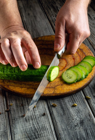 Hands skillfully slice a fresh cucumber into thin rounds on a rustic wooden cutting board. The scene captures a vibrant kitchen atmosphere filled with natural light.の写真素材
